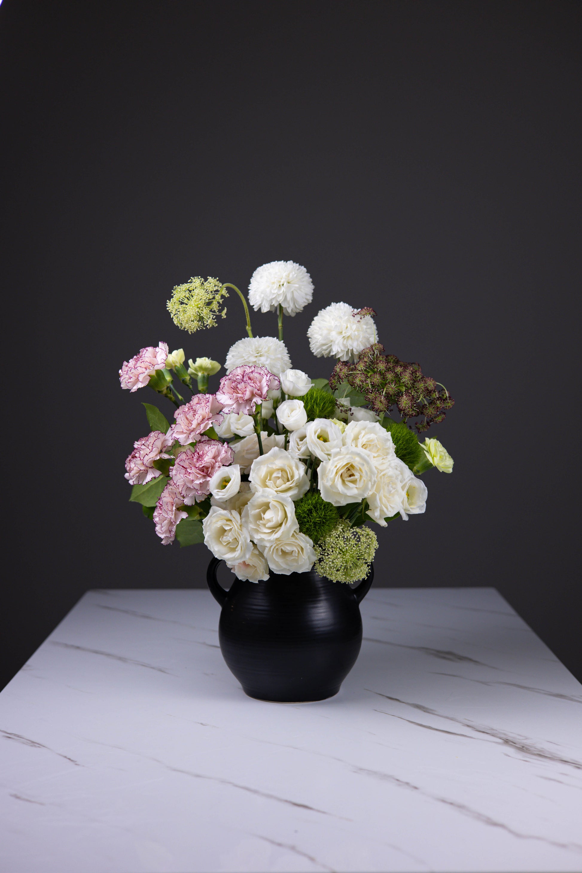 Front-facing ivory bouquet with roses, pink carnations, and green textures in a black jug vase