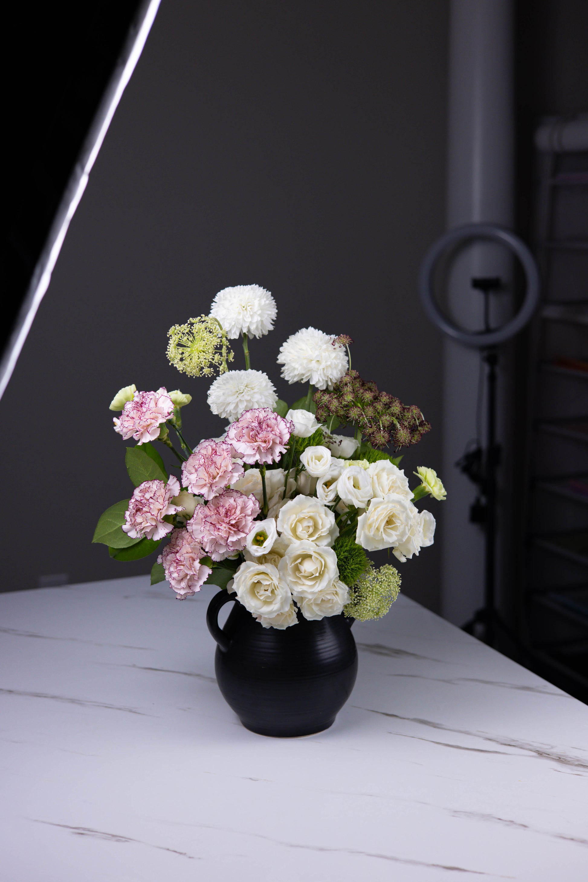 Side view of a white and pink floral bouquet styled in a modern black vase against a dark gray backdrop