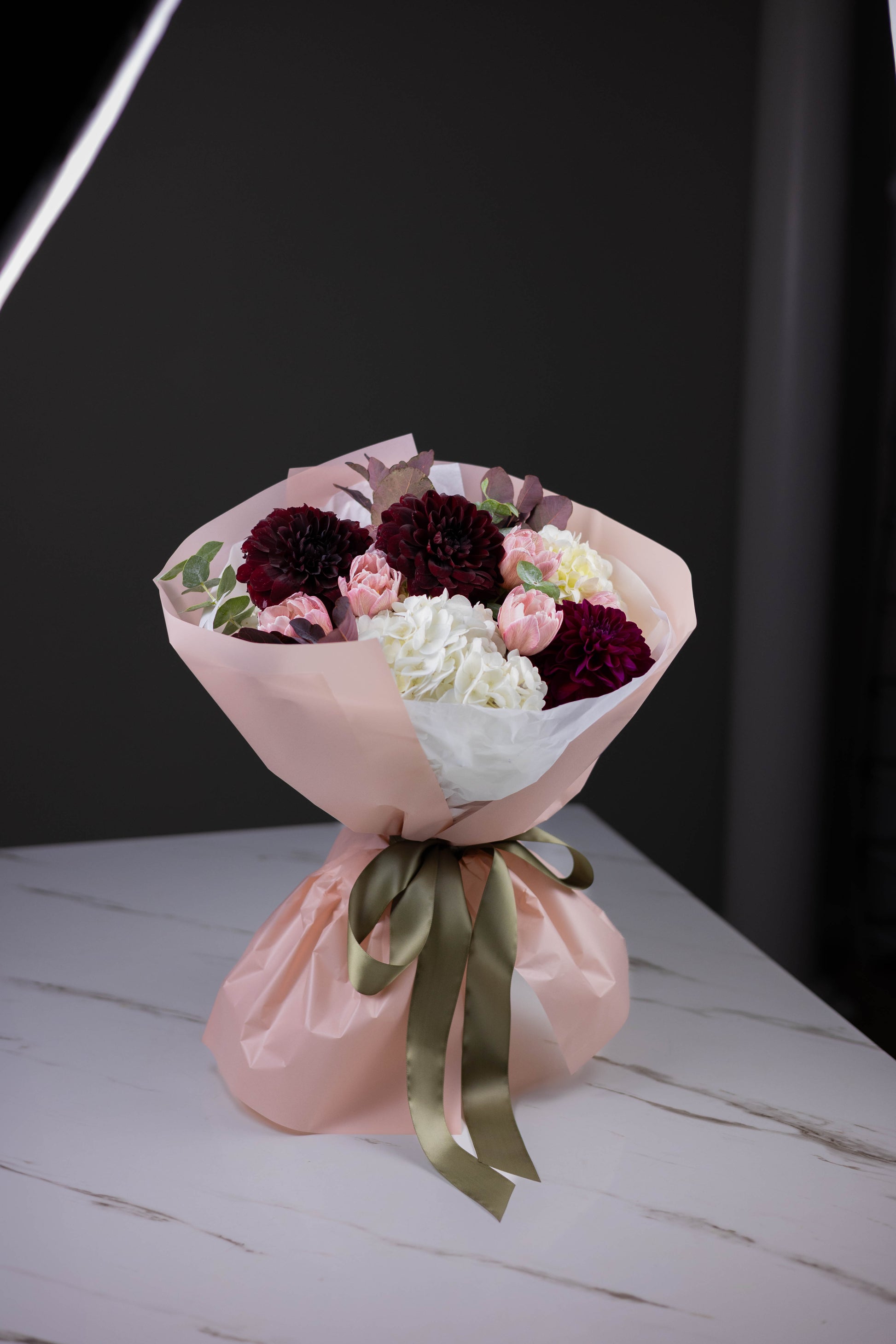 Bouquet of flowers wrapped in pink paper with a green ribbon on a white surface.