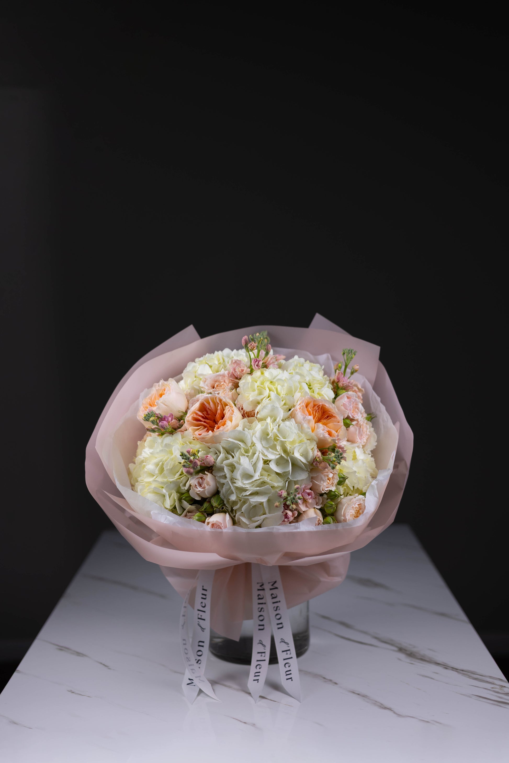 Pastel bouquet featuring white hydrangeas, blush spray roses, stock flowers, and soft peach garden roses, wrapped in pink paper with Maison d’Fleur ribbon, displayed on a white marble table.