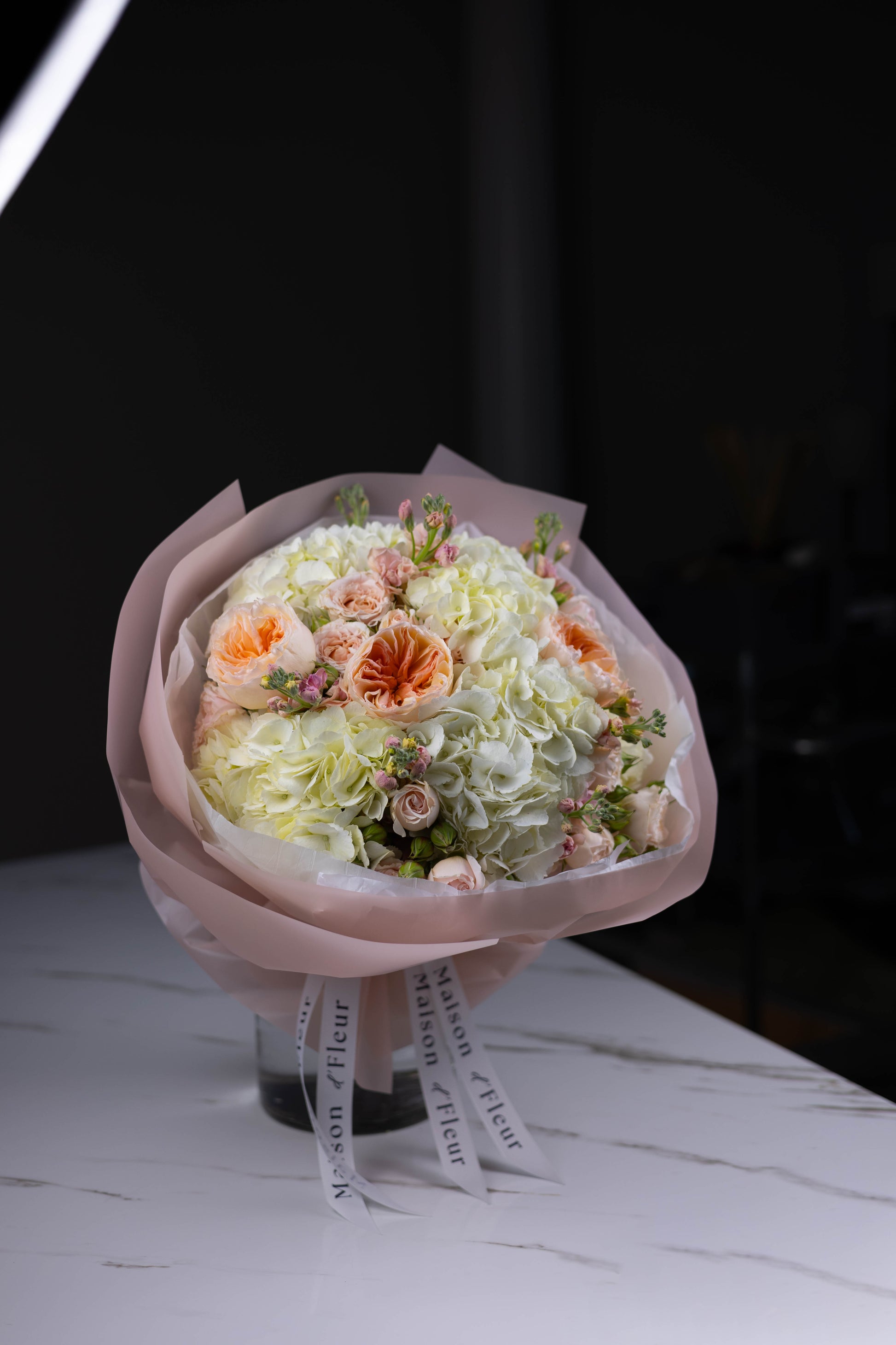 Side view of a pastel-toned bouquet with white hydrangeas, spray roses, and stock flowers, wrapped in blush paper with white branded ribbons.