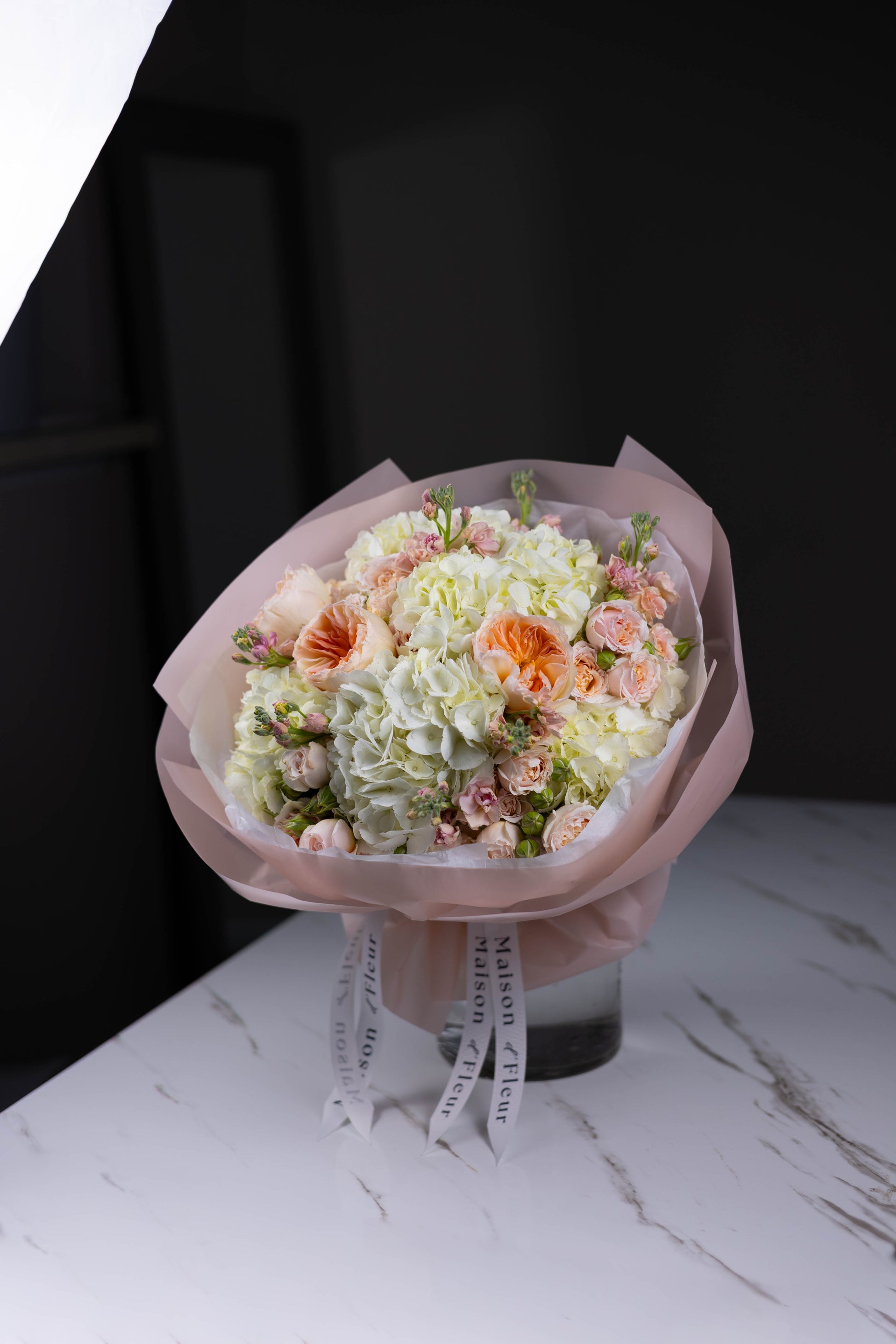 Angled shot of a round bouquet in soft pinks and creams, showcasing hydrangeas and delicate florals, standing on a marble surface.