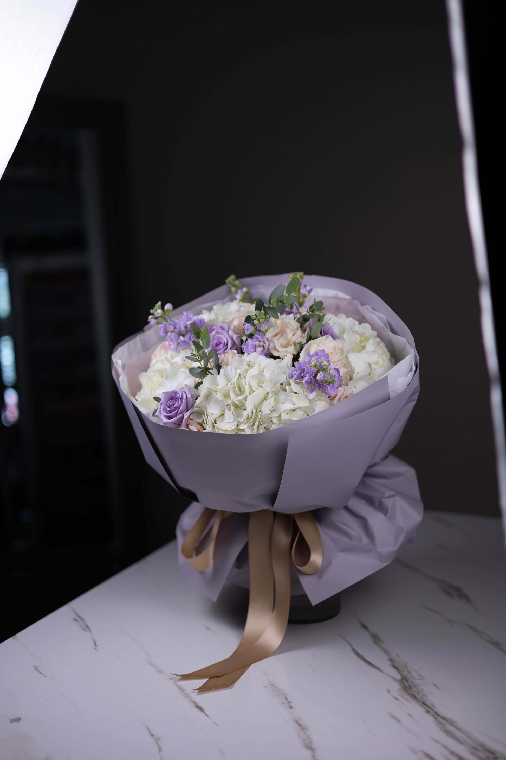 Bouquet of flowers wrapped in purple paper with a gold ribbon on a marble surface.