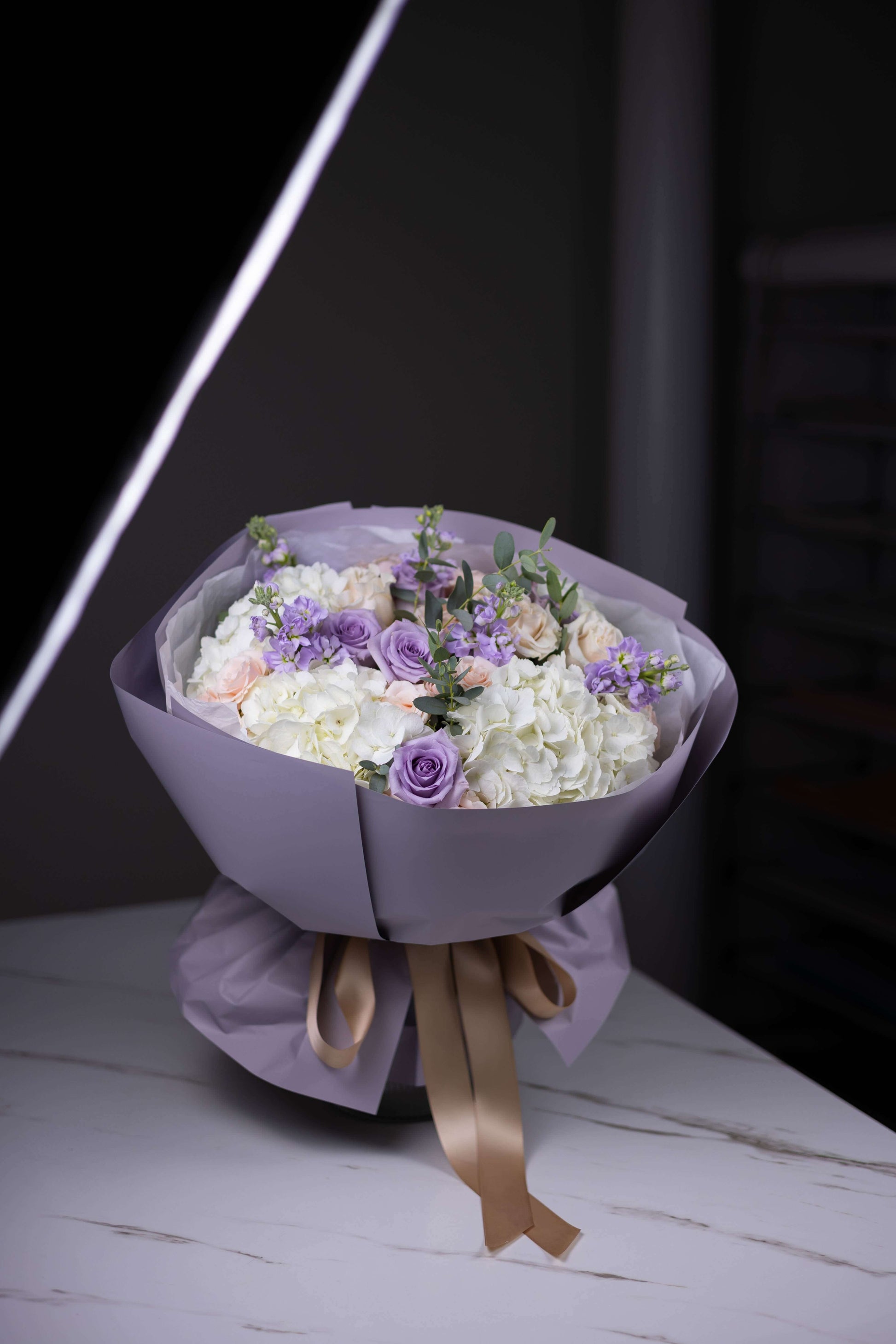 Bouquet of flowers wrapped in purple paper with a brown ribbon on a white surface.