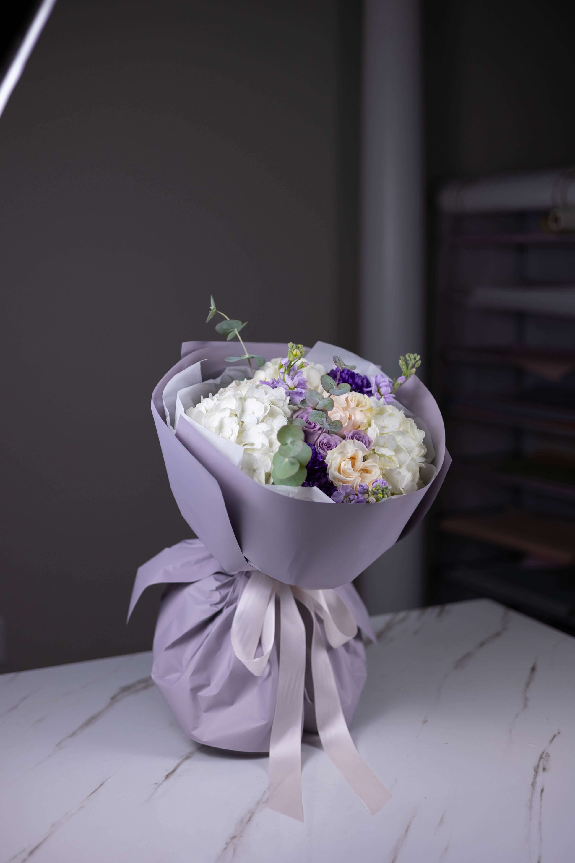 Bouquet of flowers wrapped in purple paper with a white ribbon on a marble surface.