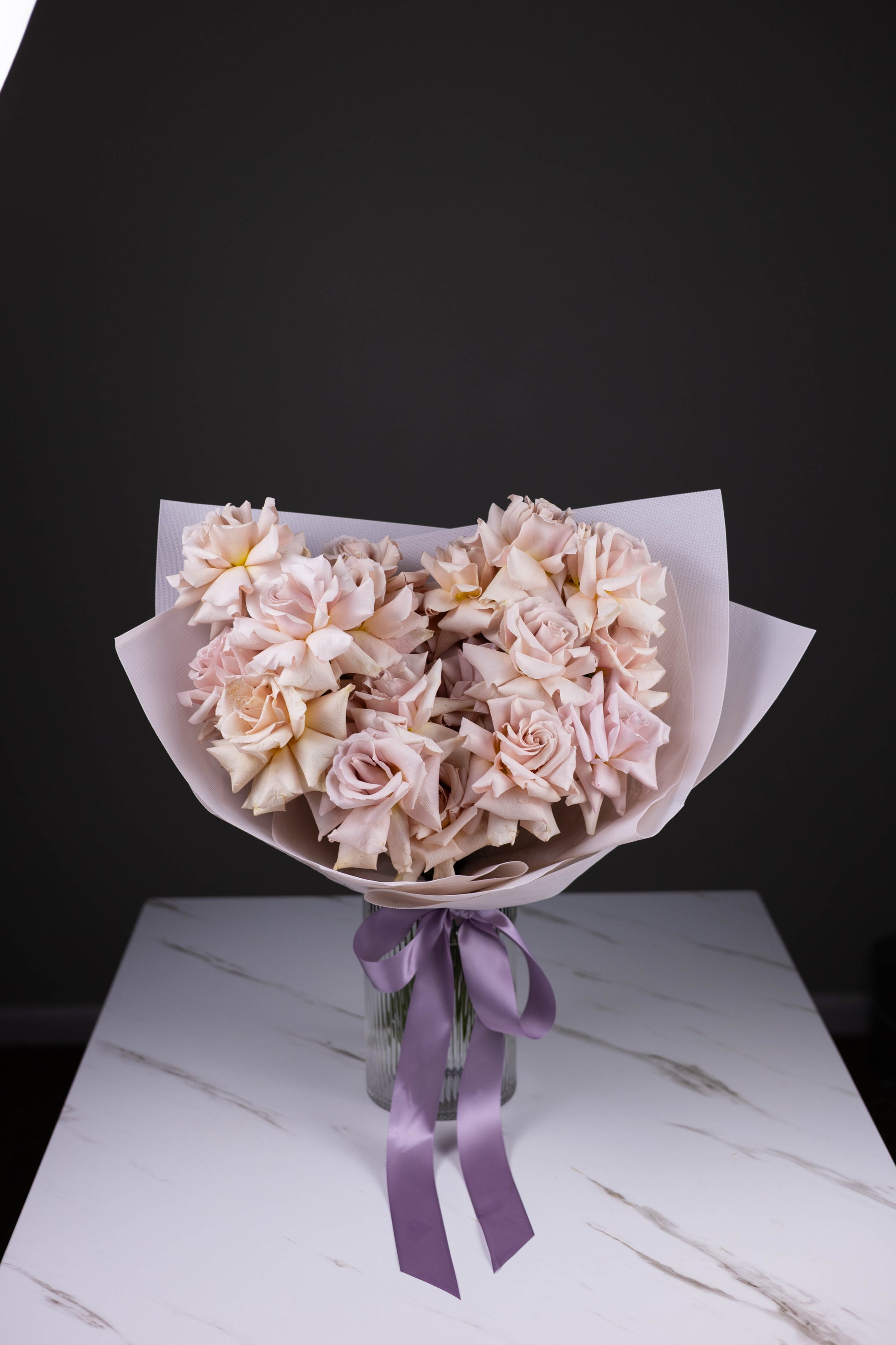 A hand-tied bouquet of blush-lavender roses in pastel wrap, displayed in a fluted glass vase with a lavender satin ribbon