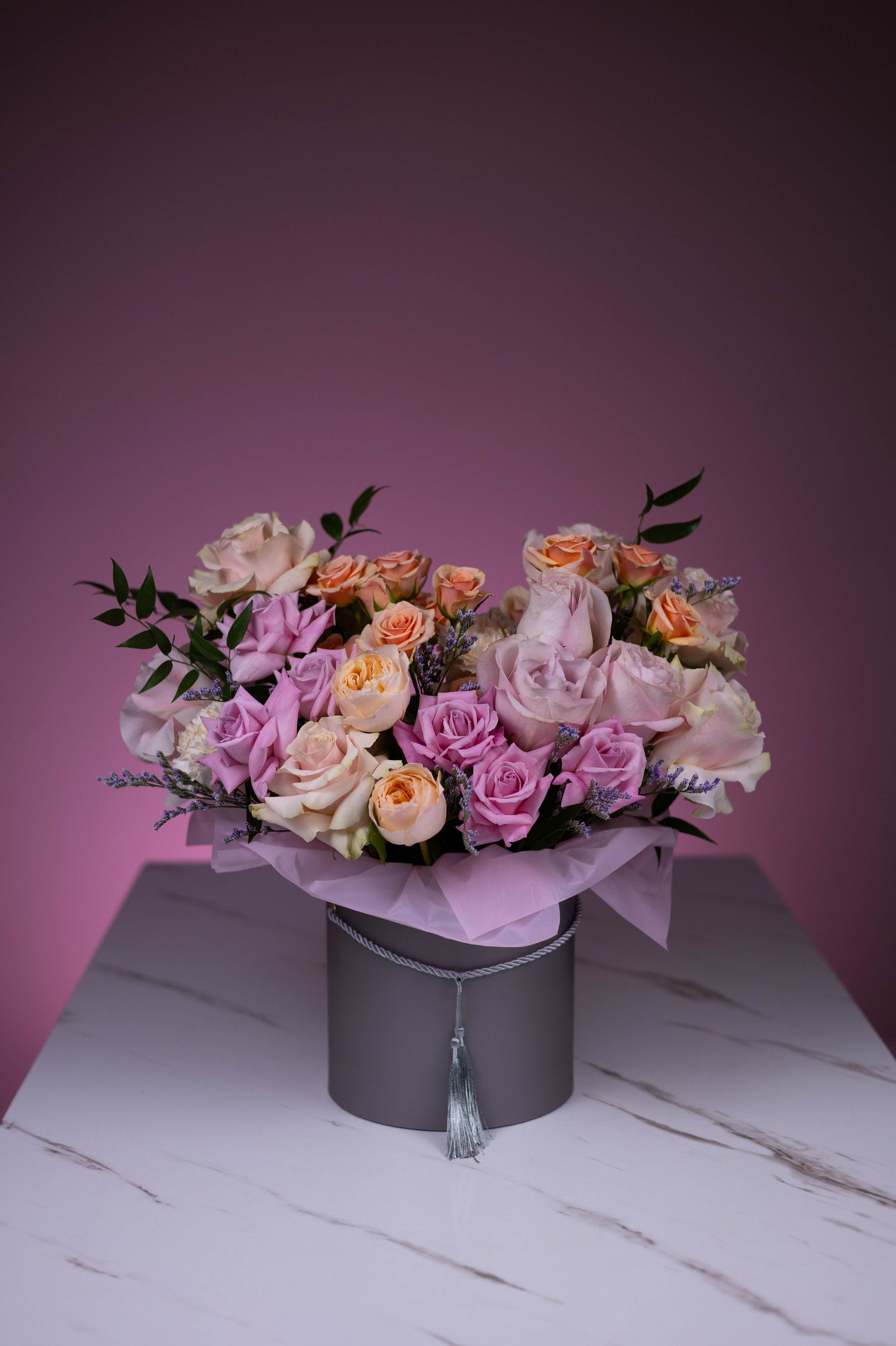 Full view of a lush round bouquet of lilac, blush, and peach roses in a Maison d’Fleur velvet grey hatbox with tassel