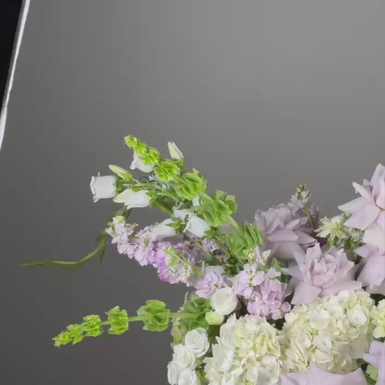 A wide-angle shot of a pastel garden arrangement featuring lavender roses, ivory hydrangeas, and soft greenery in a white compote vase.