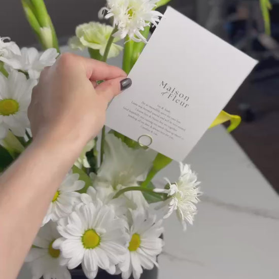 A short video showcasing a fresh spring bouquet of white daisies, yellow calla lilies, and green carnations arranged in a black vase with a Maison d’Fleur card. The camera highlights the details and textures of the flowers.