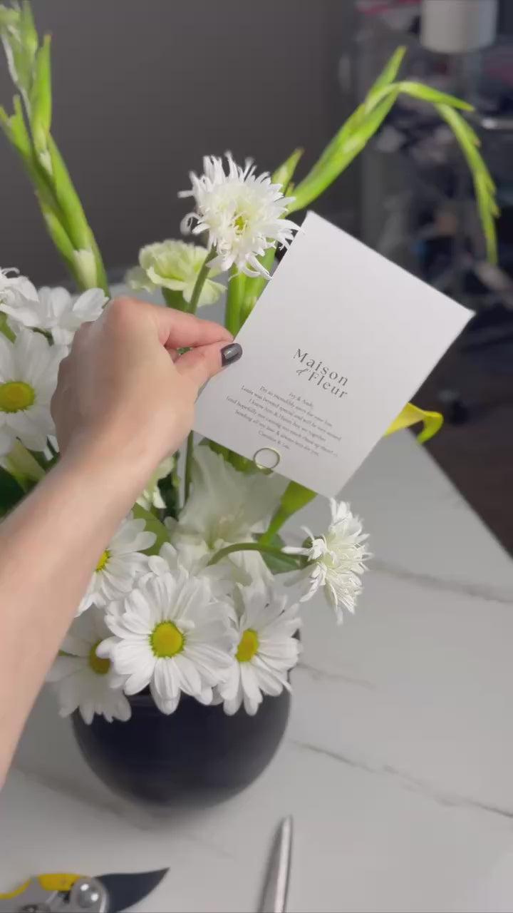 A short video showcasing a fresh spring bouquet of white daisies, yellow calla lilies, and green carnations arranged in a black vase with a Maison d’Fleur card. The camera highlights the details and textures of the flowers.